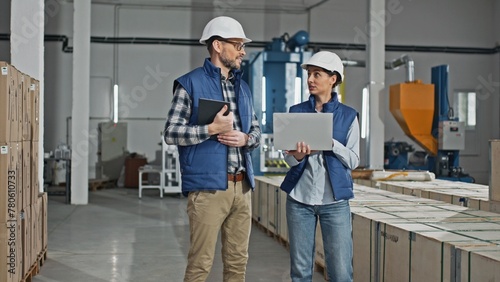 Two Caucasian workers wearing uniforms and helmets while standing in large storage or industrial facility. People actively talking together about distributing production. Using technology devices. © VAKSMANV
