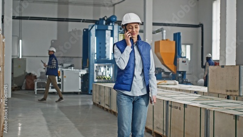 Beautiful Caucasian manager in helmet holding laptop while speaking with someone on smartphone. Young woman remotely talking with partners or clients while standing in manufacturing facility.