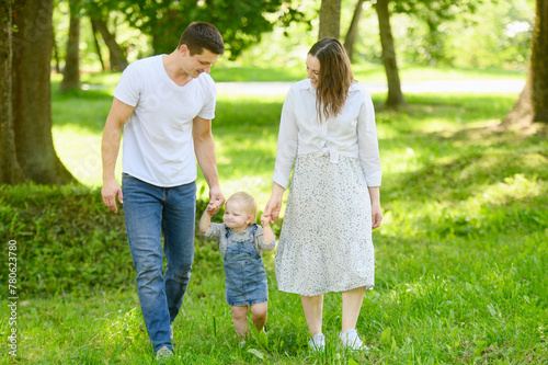 Toddler takes his first steps holding hands mom and dad in city park in the summer. Child learns walk independently using hands of his parents. Help with walking, motor skills and outdoor activities