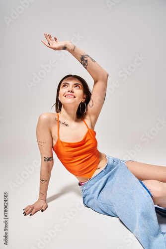 A young woman with brunette hair elegantly dressed in an orange top and blue skirt in a studio setting.