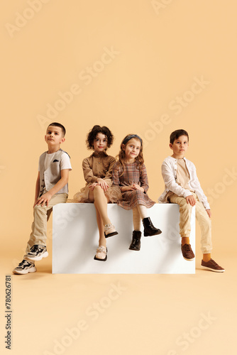 Little children, boys and girls dressed glamour outfits with trendy hairstyles posing sitting on white cube against beige background. Concept of fashion and style, beauty, childhood, Black Friday, .