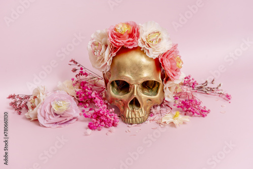 Fototapeta Naklejka Na Ścianę i Meble -  Front image of a golden skull surrounded by dried flowers on a soft pink surface