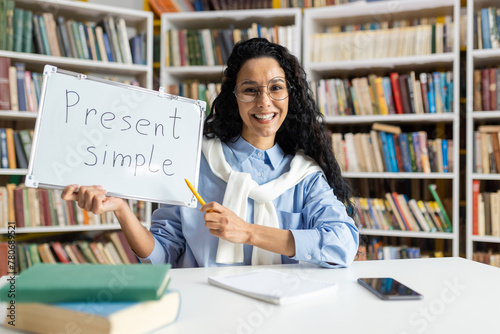 Cheerful educator teaching the present simple tense, using a whiteboard in front of a bookshelf-filled library.