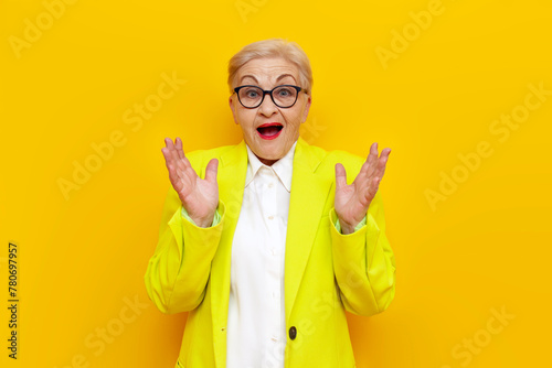 surprised old businesswoman in glasses and formal wear looks away in shock on yellow isolated background, elderly pensioner grandmother in blazer worries in amazement photo