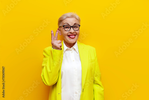 old businesswoman in glasses and formal wear showing peace gesture and tongue out on yellow isolated background, elderly pensioner grandmother in blazer showing number two with fingers photo