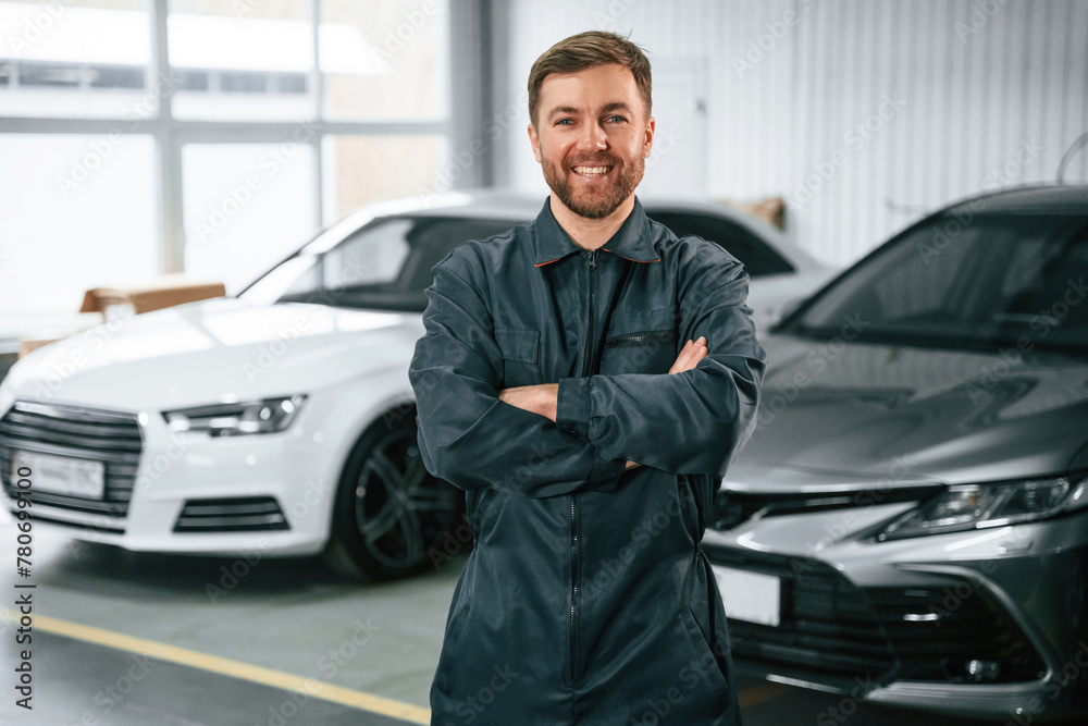 Handsome friendly guy. Repairman in uniform is standing in the garage with cars Handsome friendly guy. Repairman in uniform is standing in the garage with cars