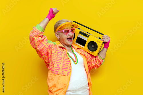 funny crazy granny in hipster clothes listening to music on tape recorder and singing on yellow isolated background, elderly cool woman dancing at party and screaming