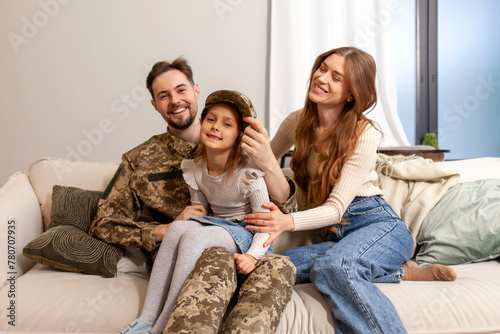Ukrainian army soldier in camouflage uniform returned home to his family, military cadet sits on the sofa with his wife and daughter and smiles, child hugs veterans father