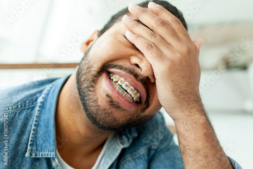 happy african american man with braces closes his eyes with his hand and shows facepalm in a white cafe, the man is embarrassed by a mistake and smiles