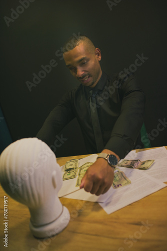 An African-American man in a shirt and tie is strangling the head of a mannequin. There is money, papers, documents on the table. Concept business, competition, stress
