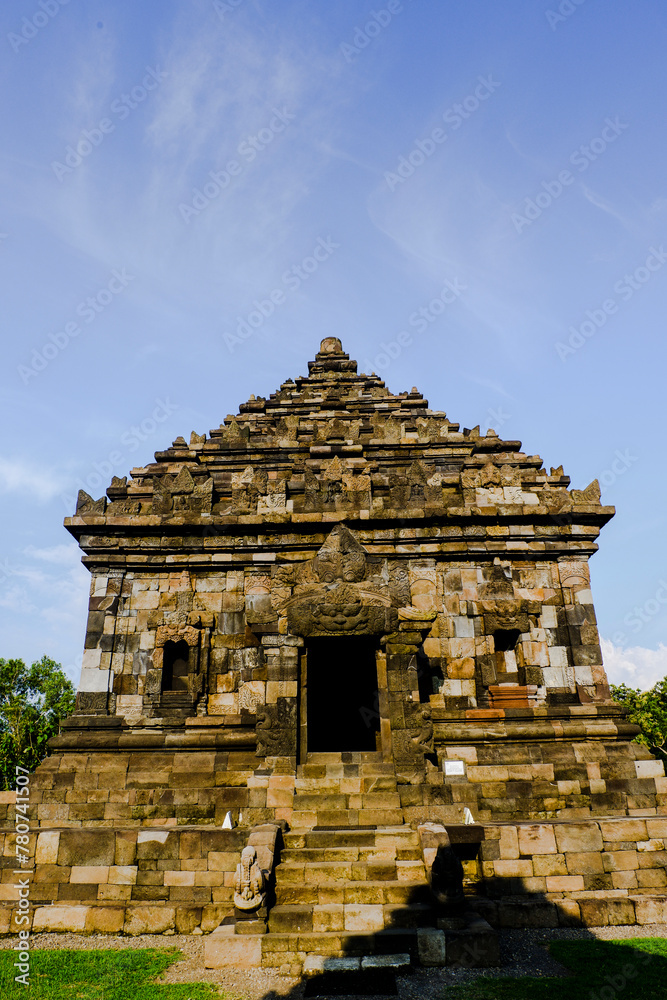 A large stone temple with a pyramidal roof, surrounded by lush green grass. The temple appears to be an ancient Hindu or Buddhist structure with architectural details.