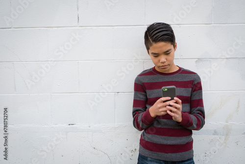 Sad teenage asian boy standing alone against outdoor grungy white background using his smartphone