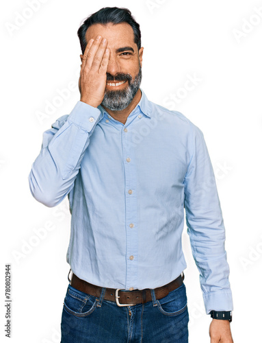 Middle aged man with beard wearing business shirt covering one eye with hand, confident smile on face and surprise emotion.