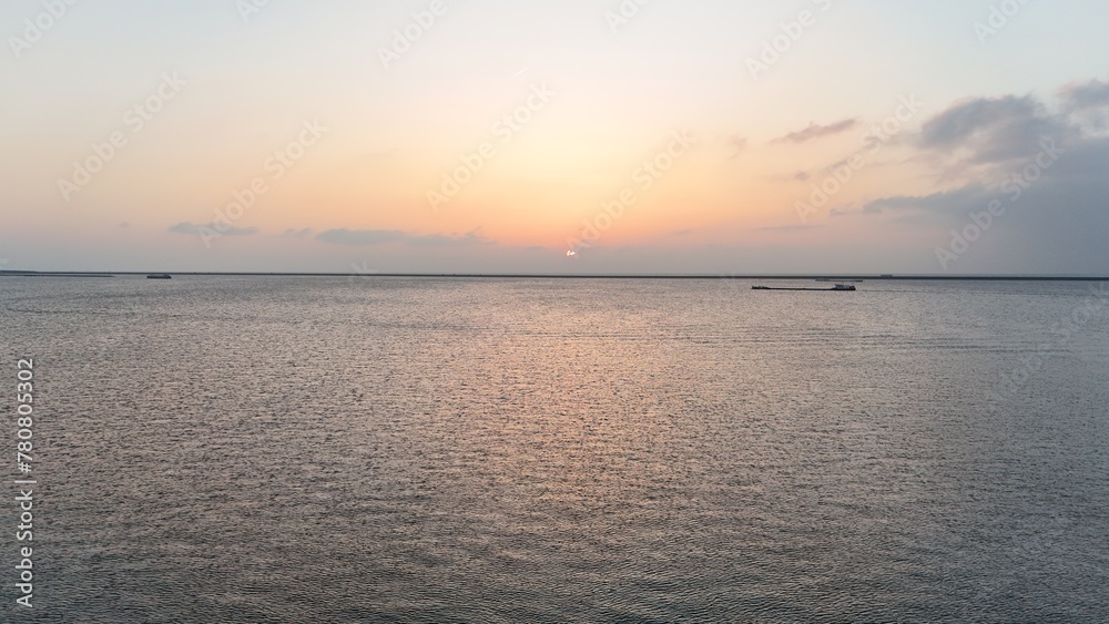 Obraz premium Tranquil seascape at sunset with gentle waves and a soft orange glow on the horizon, featuring distant cargo ships under a vast, pastel sky.