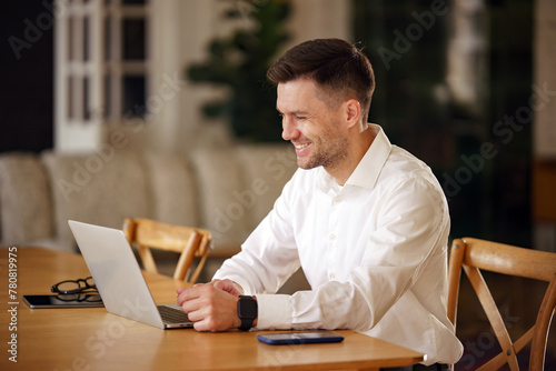 A professional gentleman smiles subtly as he interacts with a laptop at a polished wooden table, exuding an air of casual efficiency.