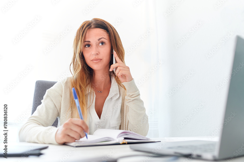 Portrait of a young entrepreneur woman talking on her cell phone and holding a pen to schedule a meeting in her agenda in the office of her workplace in a company. Portrait of a young entrepreneur woman talking on her cell phone and holding a pen to schedule a meeting in her agenda in the office of her workplace in a company.