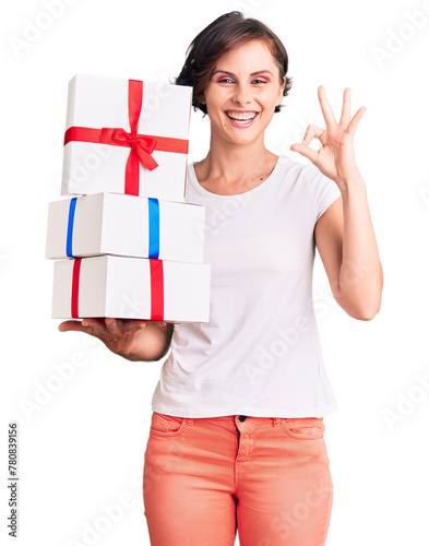 Beautiful young woman with short hair holding gifts doing ok sign with fingers, smiling friendly gesturing excellent symbol
