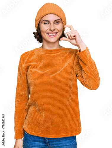 Young hispanic woman wearing casual clothes and wool cap smiling and confident gesturing with hand doing small size sign with fingers looking and the camera. measure concept.