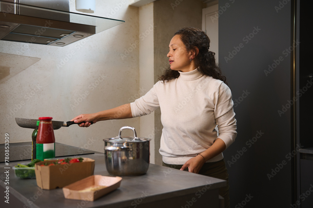 Happy pleasant woman, housewife holding a frying pan above an electric stove, cooking dinner standing at kitchen island in the home kitchen Happy pleasant woman, housewife holding a frying pan above an electric stove, cooking dinner standing at kitchen island in the home kitchen
