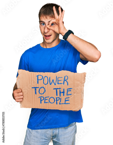 Young blond man holding power to the people banner smiling happy doing ok sign with hand on eye looking through fingers