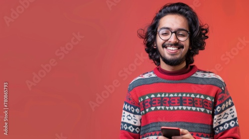 A cheerful young Indian man in a festive sweater using a smartphone on a red background. The concept of festive fun and digital communication. Design for seasonal marketing