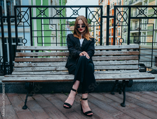 A woman in sunglasses, a white shirt and a black jacket is sitting on a wooden bench on the street in the city.