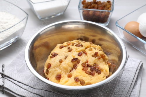 Dough with raisins in bowl and ingredients on white table, closeup