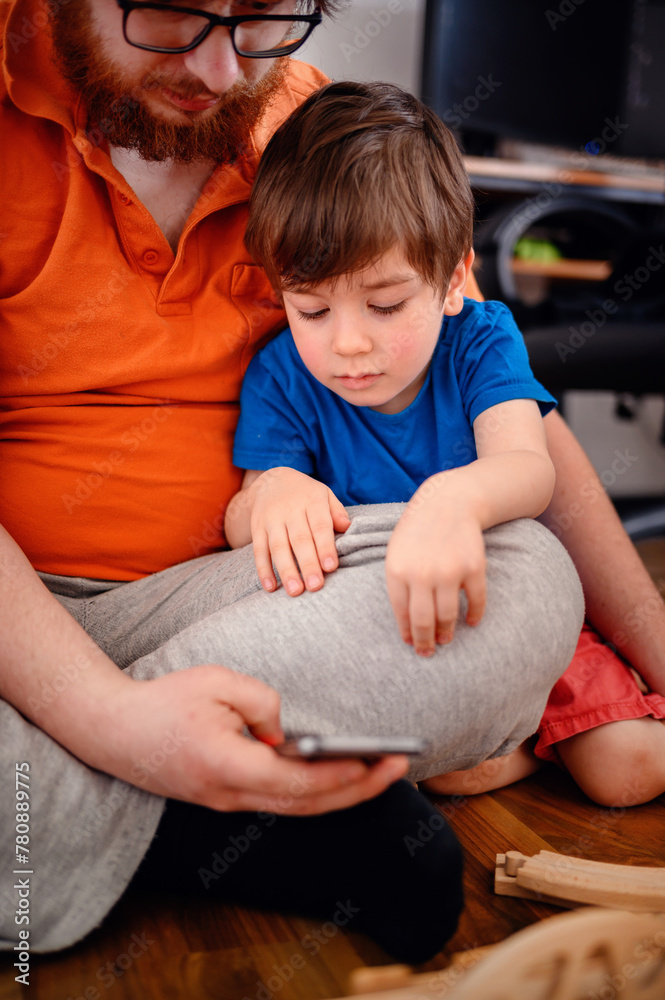 Fototapeta premium An adult and a young child explore a smartphone together, mixing the digital world with quality family time on the playroom floor