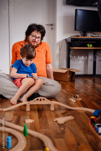 Laughing together, a child and an adult find joy in both a smartphone and a classic wooden train set, embodying the warmth of family playtime
