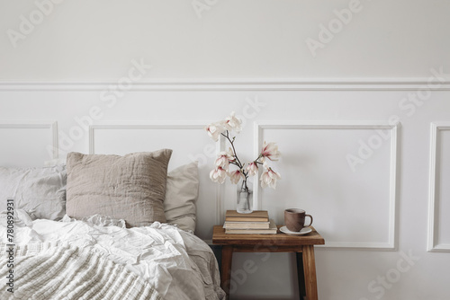 Elegant bedroom. Wooden night stand with fluted glass vase. Boolming magnolia tree branches. Cup of coffee on old books. Scandinavian interior. Linen bedding. White wall background, stucco decor.