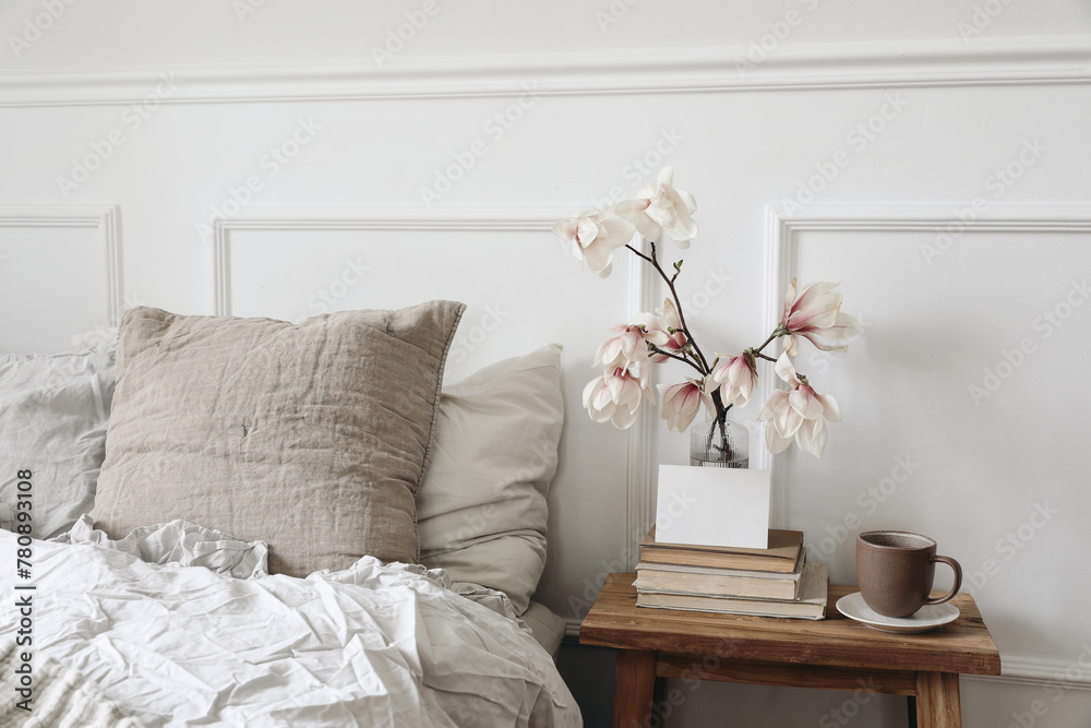 Fototapeta premium Blank greeting card, invitation mockup on old books. Elegant bedroom. Linen pillows, blanket. Wooden night stand, rippled glass vase. Blooming magnolia branches, cup of coffee. Scandinavian interior.