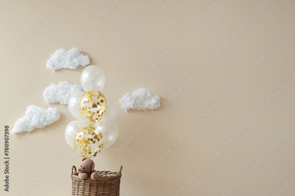 Basket with toy bear, balloons and cotton clouds on light background