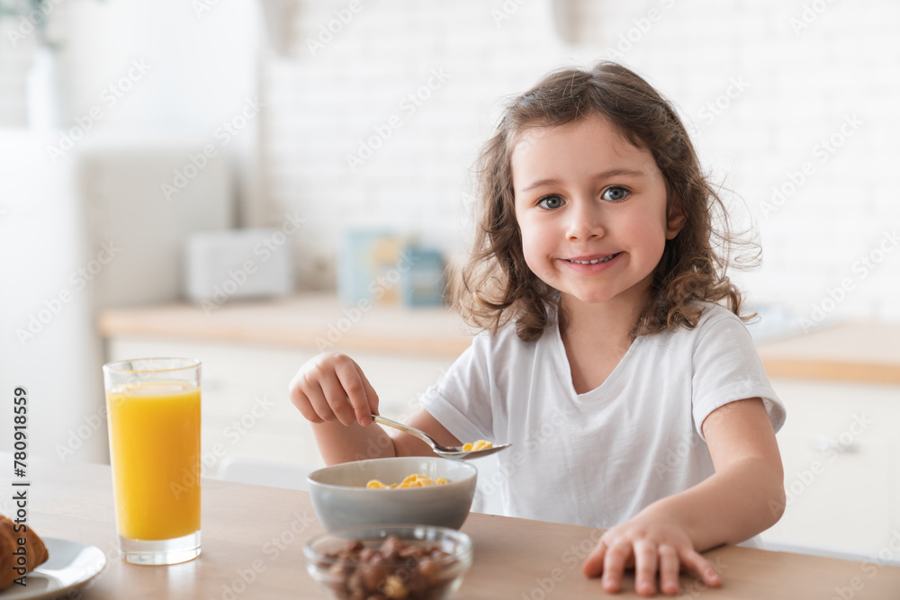 Cheerful little caucasian girl daughter having breakfast, eating corn flakes, drinking orange juice in the kitchen in the morning looking at the camera. Healthy eating habits, learning how to eat