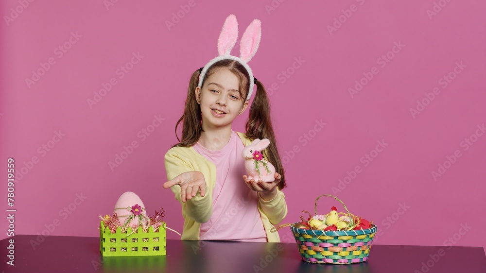 Fototapeta premium Young schoolgirl blowing air kisses in front of camera while she creates festive lovely arrangements on stuffed rabbit toy. Cheerful young child being excited about easter celebration. Camera A.