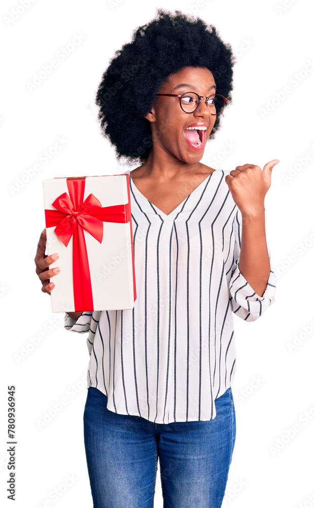 Young african american woman holding gift pointing thumb up to the side smiling happy with open mouth Young african american woman holding gift pointing thumb up to the side smiling happy with open mouth