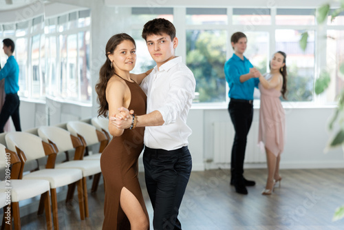 Stylish young couple, guy in white shirt and attractive girl in fitted brown dress rehearsing elegant waltz in spacious choreographic studio