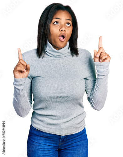 Beautiful hispanic woman wearing turtleneck sweater amazed and surprised looking up and pointing with fingers and raised arms.