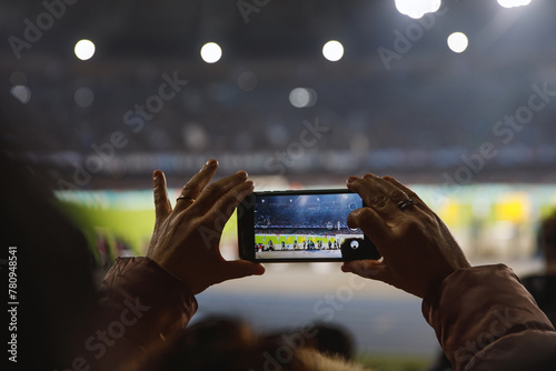 A person s hands holding a smartphone, capturing a match at a brilliantly illuminated stadium