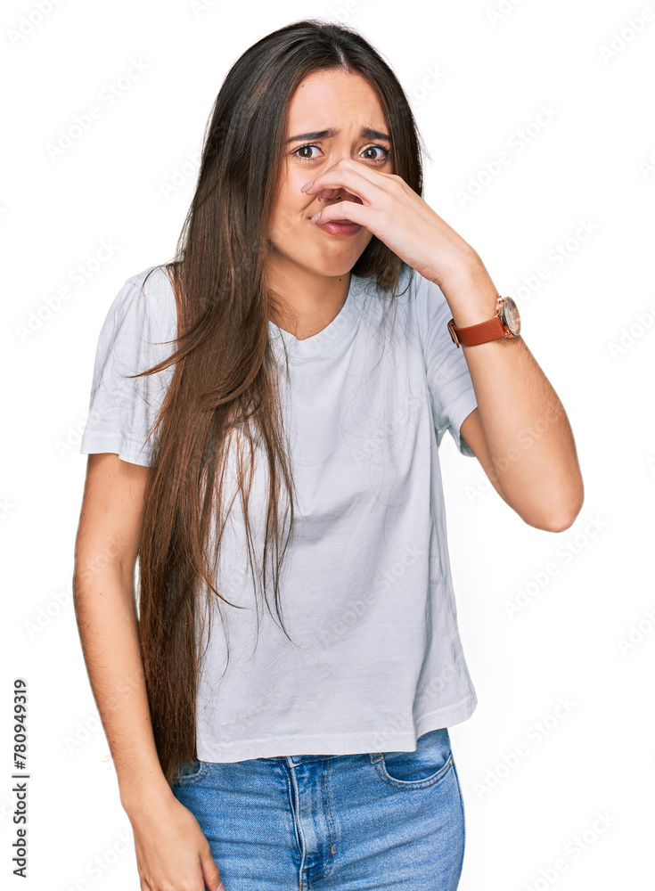 Young hispanic girl wearing casual white t shirt smelling something stinky and disgusting, intolerable smell, holding breath with fingers on nose. bad smell Young hispanic girl wearing casual white t shirt smelling something stinky and disgusting, intolerable smell, holding breath with fingers on nose. bad smell