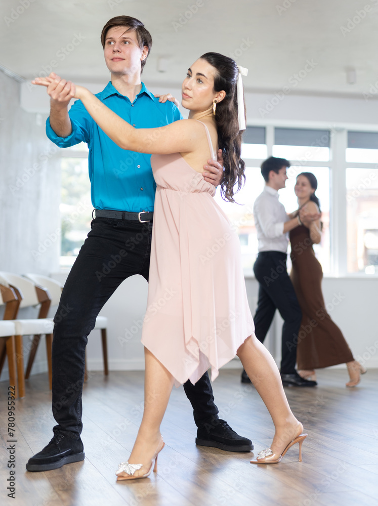 Positive emotional young guy in blue shirt enjoying impassioned merengue with female partner wearing elegant dress in latin dance class. Amateur dancing concept..