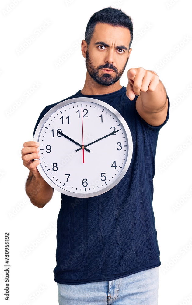 Young hispanic man holding big clock pointing with finger to the camera and to you, confident gesture looking serious Young hispanic man holding big clock pointing with finger to the camera and to you, confident gesture looking serious