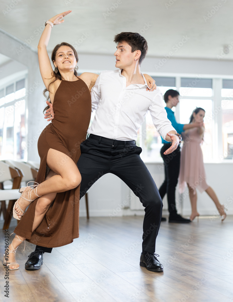 Fototapeta premium Stylish emotional young hispanic guy in white shirt and black trousers enjoying impassioned merengue with female partner wearing elegant dress in latin dance class. Amateur dancing concept