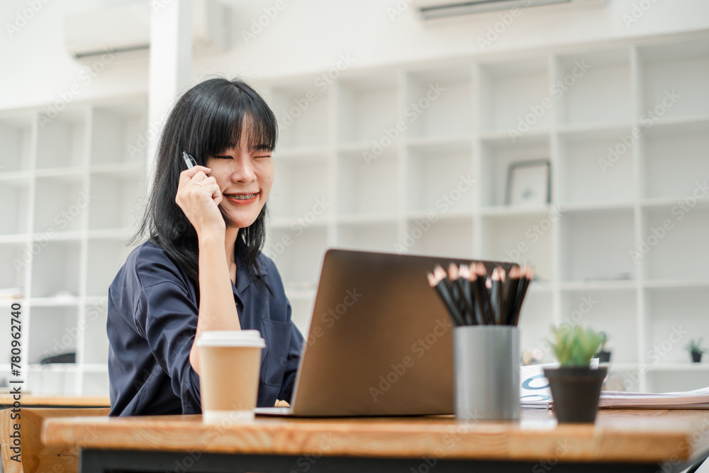 Cheerful young woman with braces talking on the phone while working on a laptop in a bright office setting. Cheerful young woman with braces talking on the phone while working on a laptop in a bright office setting.