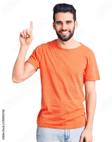 Young handsome man with beard wearing casual t-shirt showing and pointing up with finger number one while smiling confident and happy.
