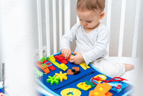 Baby sitting in a crib playing with montessori busy book. Concept of keeping children from screen by activity books and quiet books