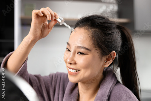 Smiling Asian Woman Using Serum For Skincare Routine At Home. Feeling Content and Fresh, Engaged In Daily Beauty Treatment With Dropper Bottle. Personal Care and Wellbeing Concept.