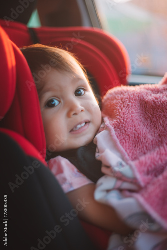 Asian Toddler Girl seats peacefully in her red carseat. She is holding her pink blanket while sucking on her thumb during sunrise or sunset. While looking at the camera.