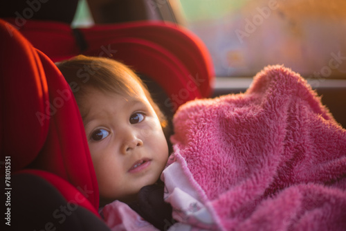 Asian Toddler Girl seats peacefully in her red carseat. She is holding her pink blanket while sucking on her thumb during sunrise or sunset. While looking at the camera.