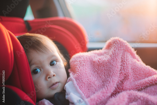 Asian Toddler Girl seats peacefully in her red carseat. She is holding her pink blanket while sucking on her thumb during sunrise or sunset. While looking at the camera.