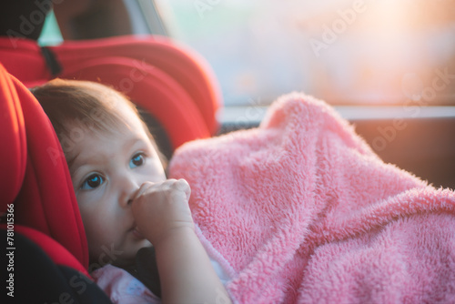 Asian Toddler Girl seats peacefully in her red carseat. She is holding her pink blanket while sucking on her thumb during sunrise or sunset. While looking at the camera.